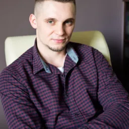 Portret młodego mężczyzny w kraciastej koszuli, siedzącego na kremowym fotelu na tle szarej ściany.