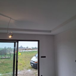 FIRMA OGÓLNOBUDOWLANA MULCZYN TYNK ŁUKASZ MULCZYŃSKI - Wykończone wnętrze z gładkimi, białymi ścianami i ozdobnym podwieszanym sufitem. Przez otwarte drzwi balkonowe widok na podwórze z samochodem.