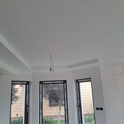 FIRMA OGÓLNOBUDOWLANA MULCZYN TYNK ŁUKASZ MULCZYŃSKI - Białe wnętrze po remoncie z trzema oknami i dekoracyjnym sufitem. Widoczne detale wykończenia i elementy instalacji elektrycznej. Świeżo malowane ściany.