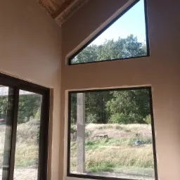 Wnętrze domu w Dolsku z oknami i surowymi ścianami po nałożeniu tynku, widoczny sufit z drewnianych belek i fragment instalacji kanalizacyjnej.