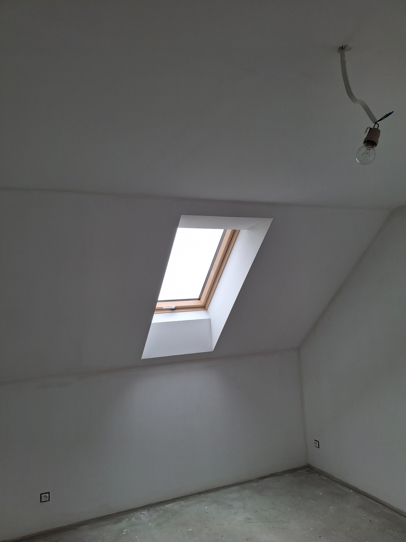 Poddasze po remoncie: białe ściany, okno dachowe z drewnianą ramą, surowa żarówka na kablu. Widoczne gniazdka elektryczne. Podłoga betonowa.