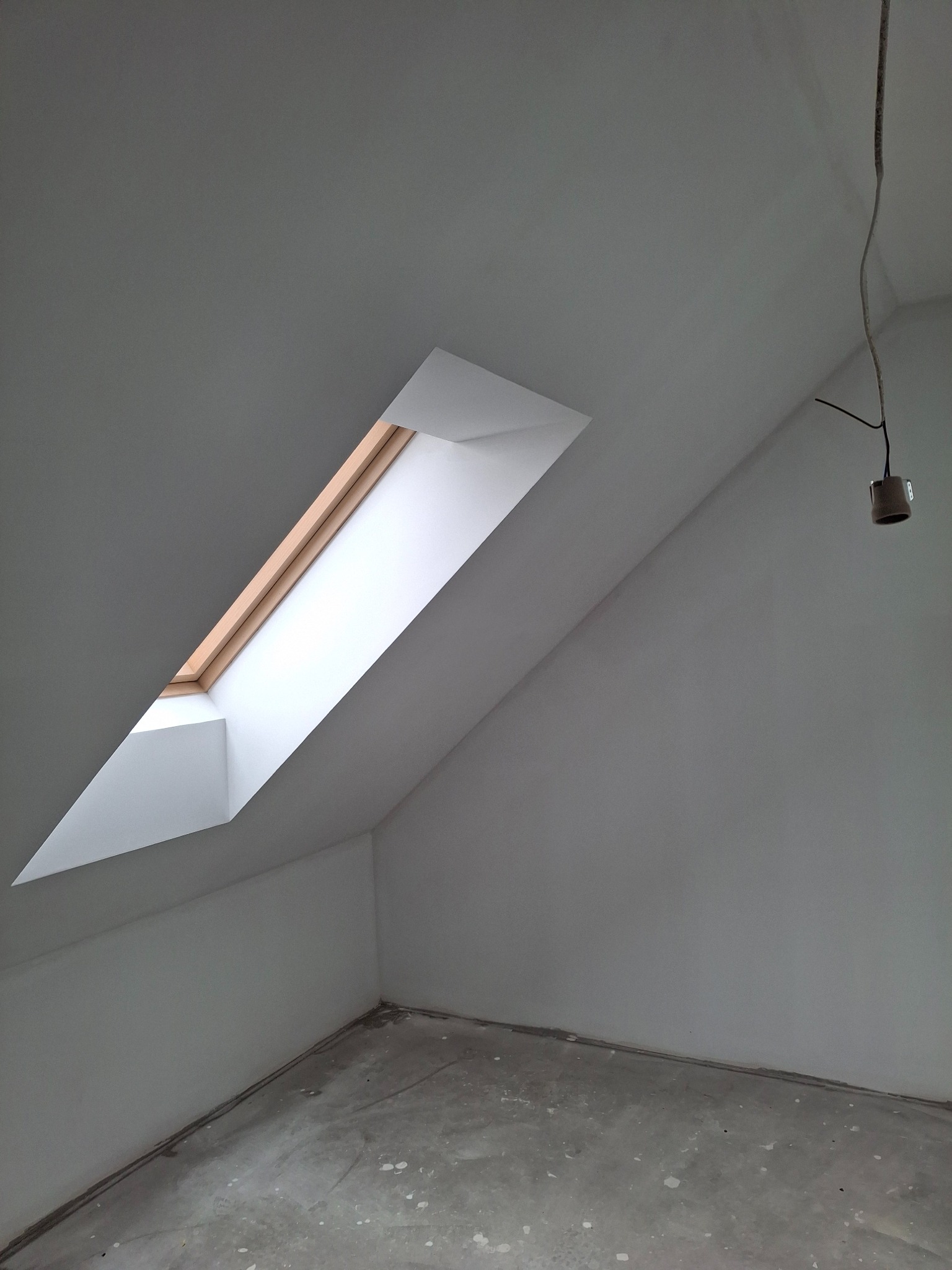 Poddasze ze świeżo wykończonymi ścianami i oknem dachowym. Widoczny surowy beton na podłodze i wisząca oprawa oświetleniowa. Stonowana kolorystyka.