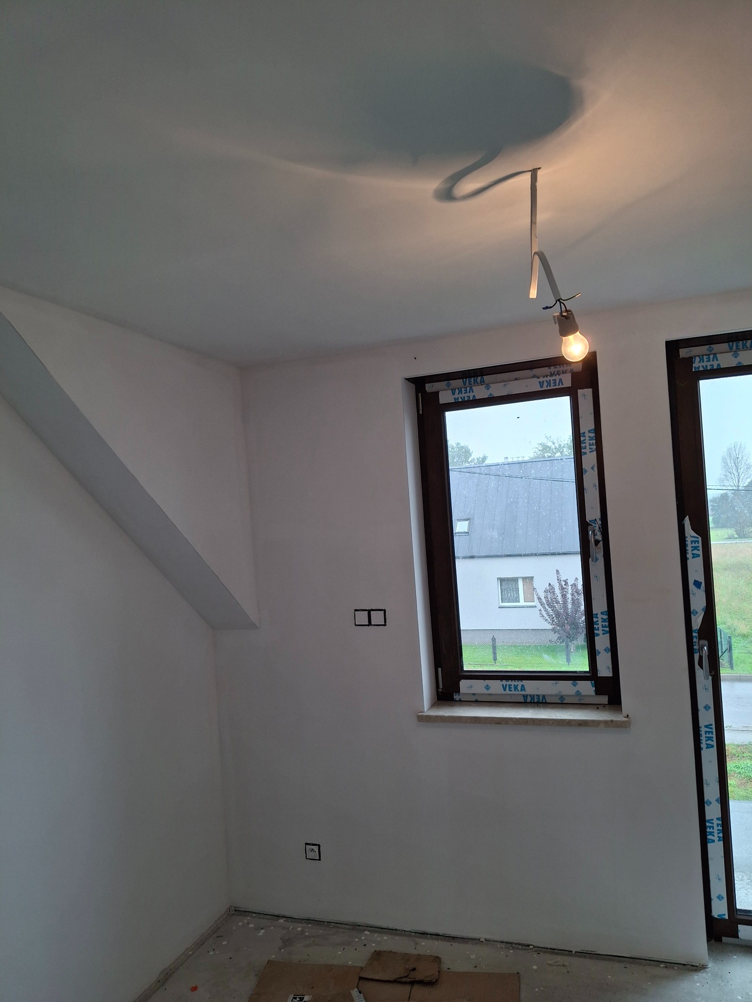 Wnętrze po szpachlowaniu: gładkie, białe ściany, dwa okna z folią ochronną VEKA, surowe oświetlenie z żarówką. Widoczne gniazdka elektryczne i poddasze.