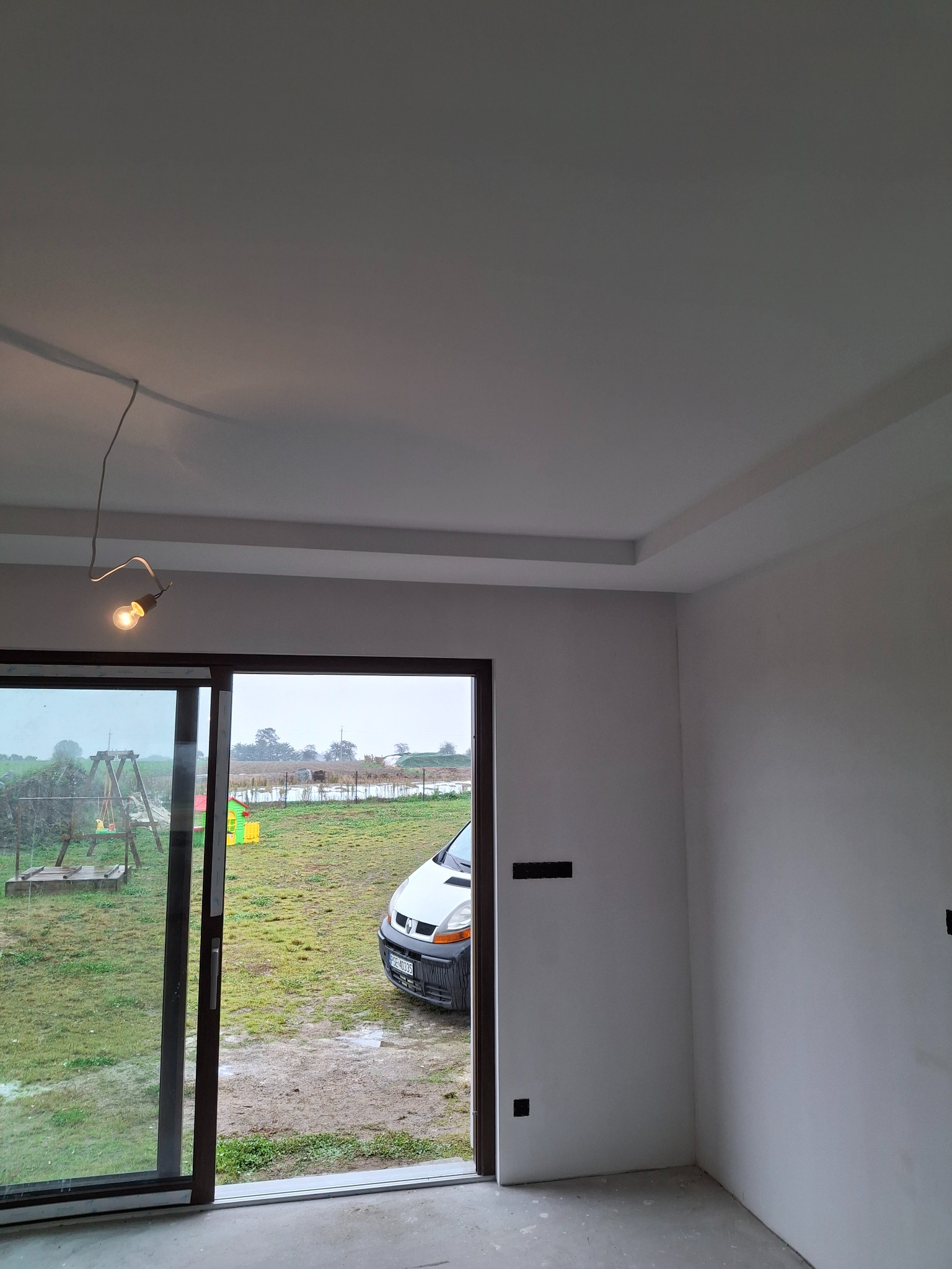 Wykończone wnętrze z gładkimi, białymi ścianami i ozdobnym podwieszanym sufitem. Przez otwarte drzwi balkonowe widok na podwórze z samochodem.