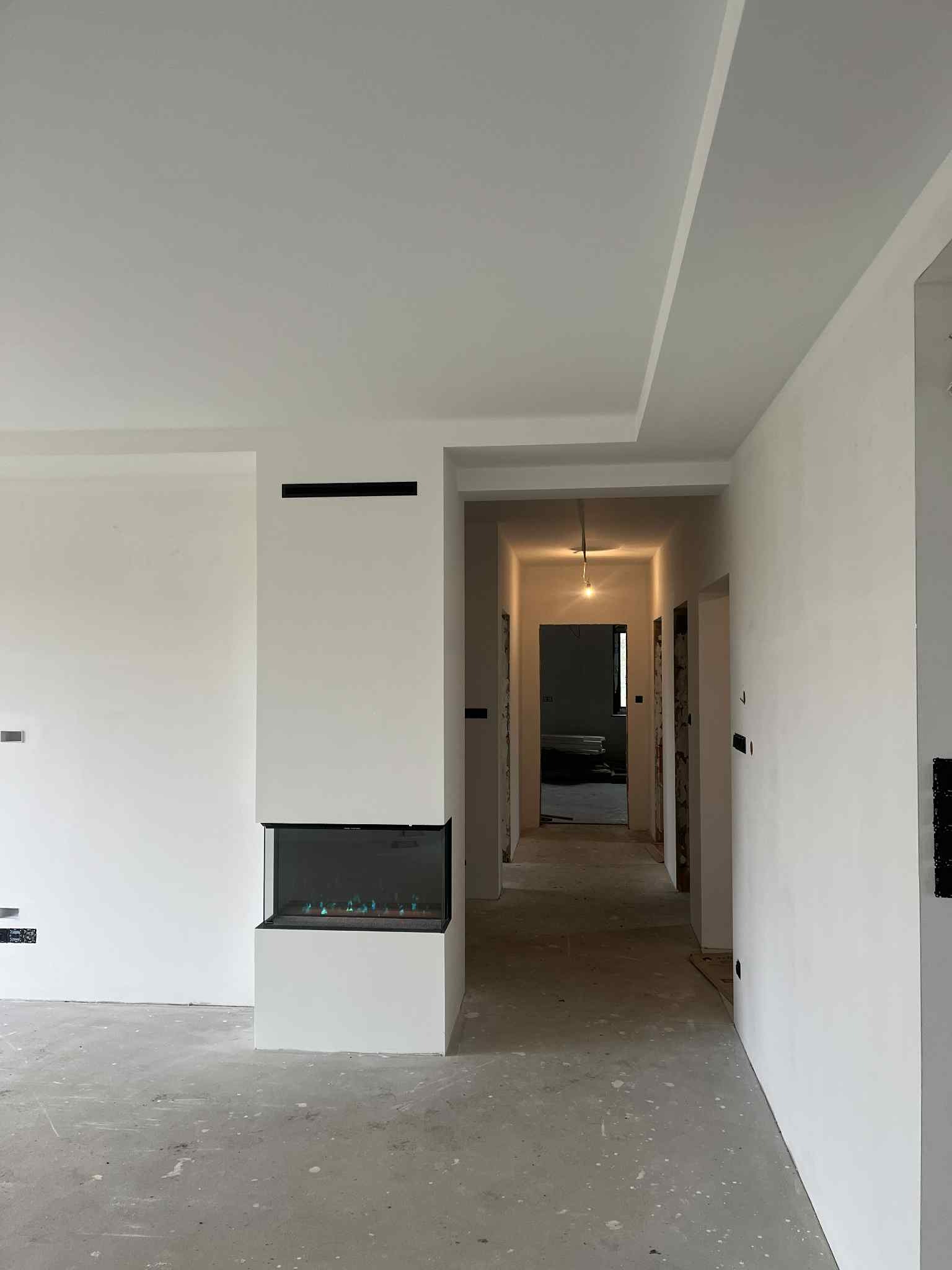 Wnętrze domu w trakcie wykańczania: białe ściany, sufit z podwieszanym elementem, narożny kominek elektryczny. Widoczne kable i surowa podłoga.