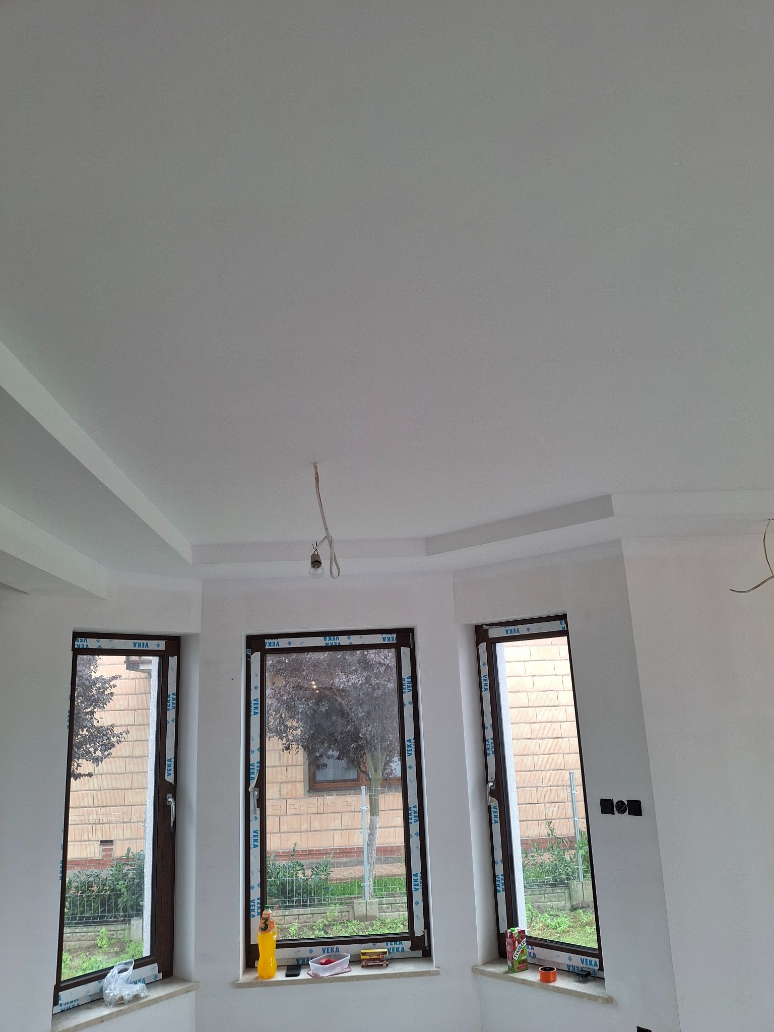 Białe wnętrze po remoncie z trzema oknami i dekoracyjnym sufitem. Widoczne detale wykończenia i elementy instalacji elektrycznej. Świeżo malowane ściany.