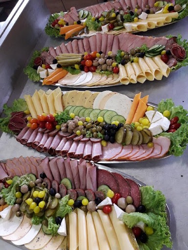 Cztery owalne patery z wędlinami, serami, warzywami i oliwkami, dekorowane sałatą, ułożone kaskadowo na metalowym stole.