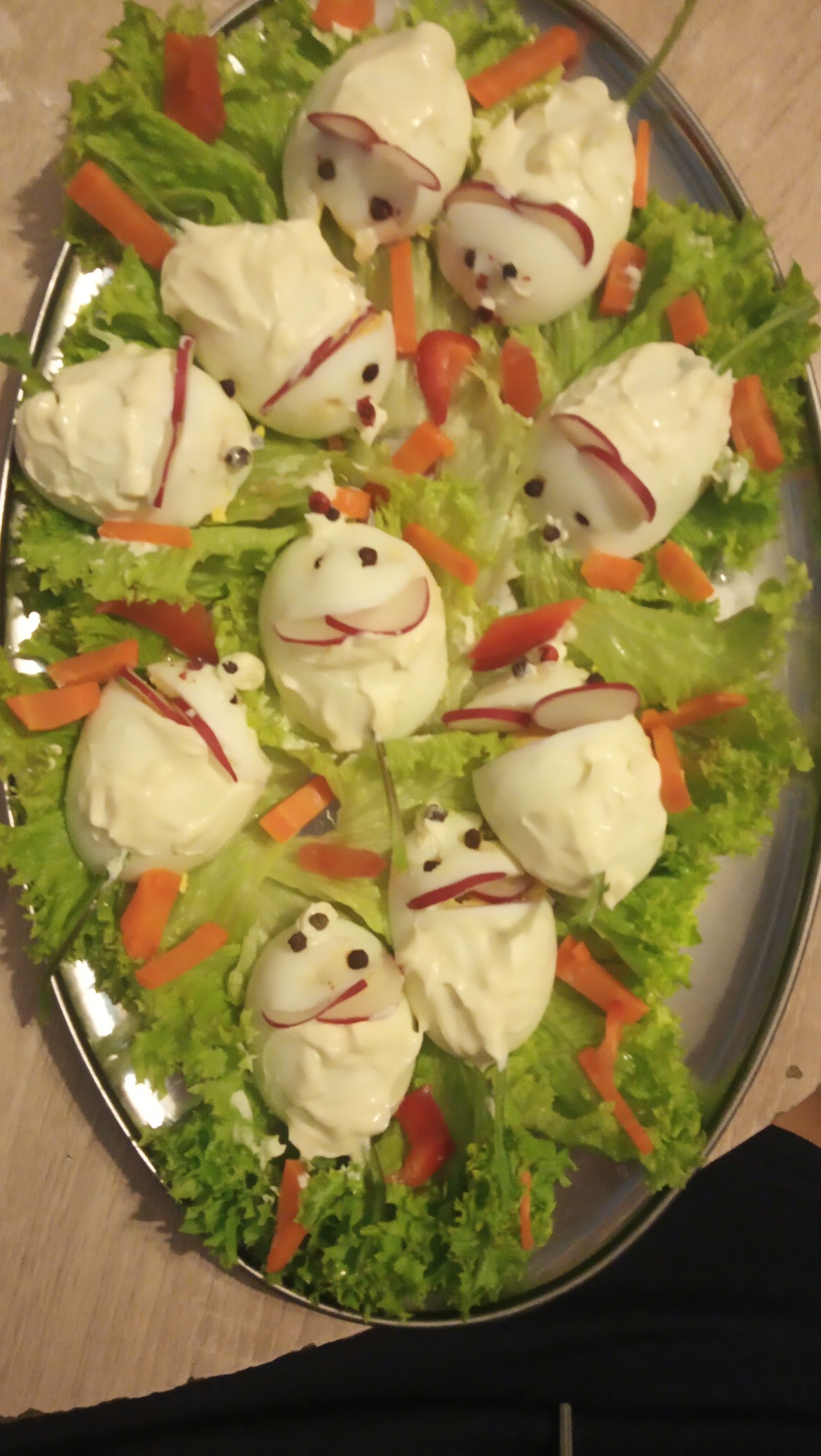 Dekoracyjne jajka w majonezie ułożone na sałacie, udekorowane rzodkiewką, marchewką i pieprzem tworzące kształty zwierząt na metalowej tacy.