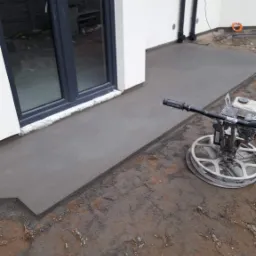 Świeżo wylana, gładka posadzka jastrychowa przy drzwiach tarasowych, obok stoi zacieraczka mechaniczna, widoczny fragment ziemi wokół wylewki.