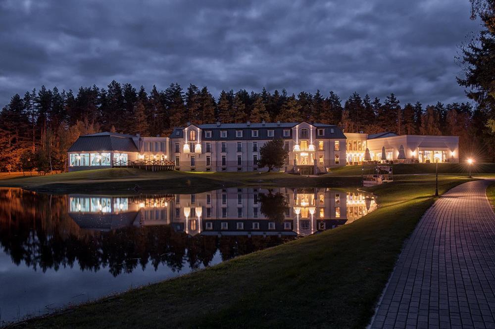 Elegancki budynek z oświetleniem zewnętrznym odbijający się w tafli jeziora w nocy, otoczony lasem i ścieżką z kostki brukowej.