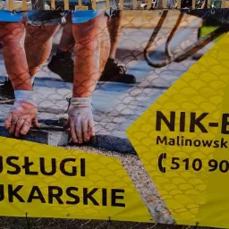 Baner reklamowy firmy NIK-BUD z numerem telefonu, prezentujący zbliżenie na dłonie w rękawicach układające szarą kostkę brukową.