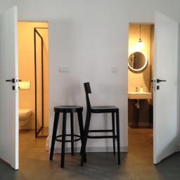 Plan A - Minimalistyczne wnętrze z otwartymi drzwiami do łazienki i toalety, czarne krzesła barowe na pierwszym planie, dominująca biel i szarość, okrągłe lustro podświetlane w tle.