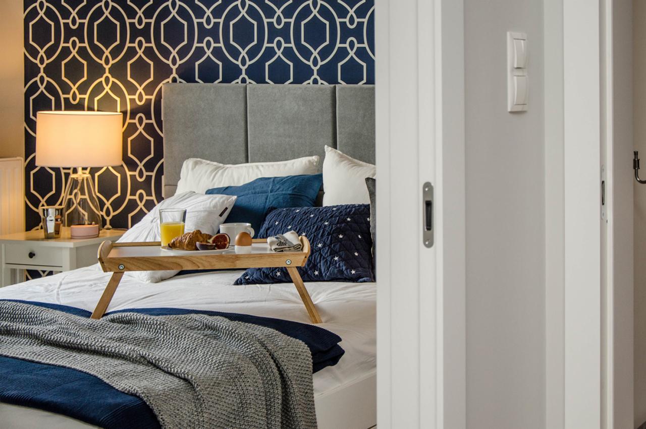 Sypialnia z łóżkiem, śniadaniem na tacy i wzorzystą tapetą w odcieniach granatu i bieli, widok zza uchylonych białych drzwi.