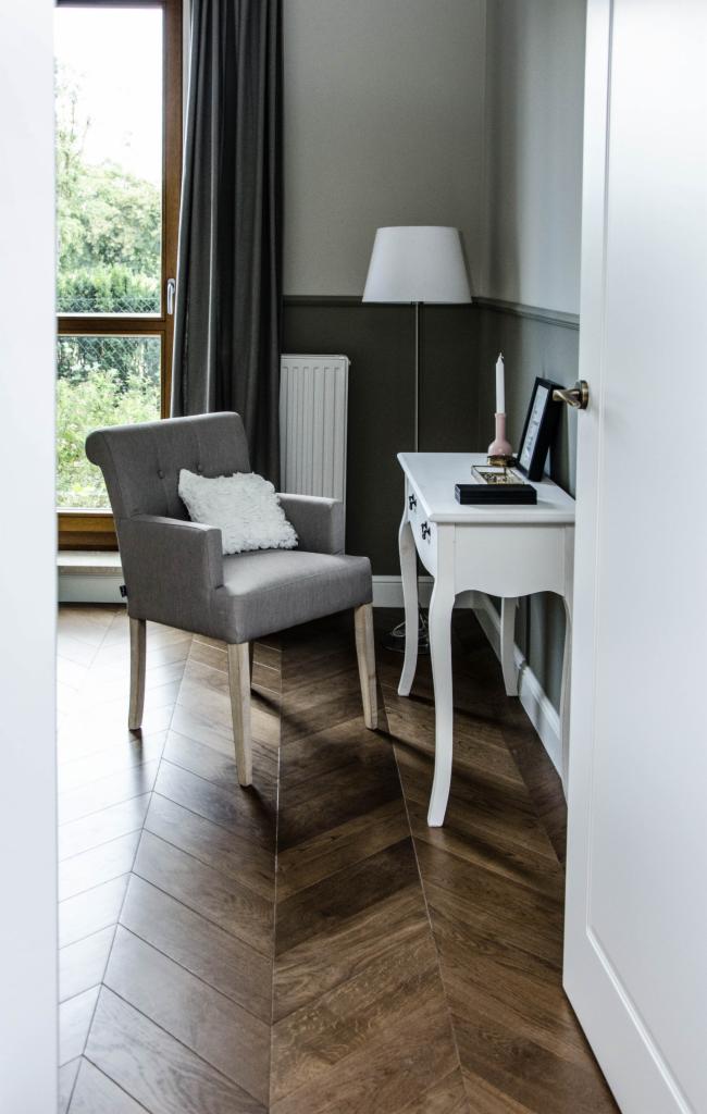 Szary fotel z poduszką przy białej toaletce na tle parkietu w jodełkę, widok zza uchylonych białych drzwi.