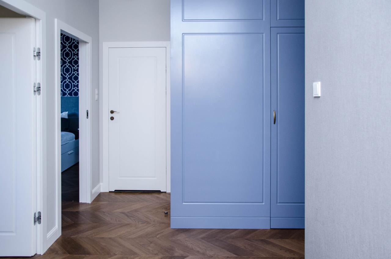 Wnętrze mieszkania z parkietem w jodełkę, białe drzwi i zabudowa szafy w odcieniach błękitu, widok na sypialnię z tapetą w geometryczny wzór.