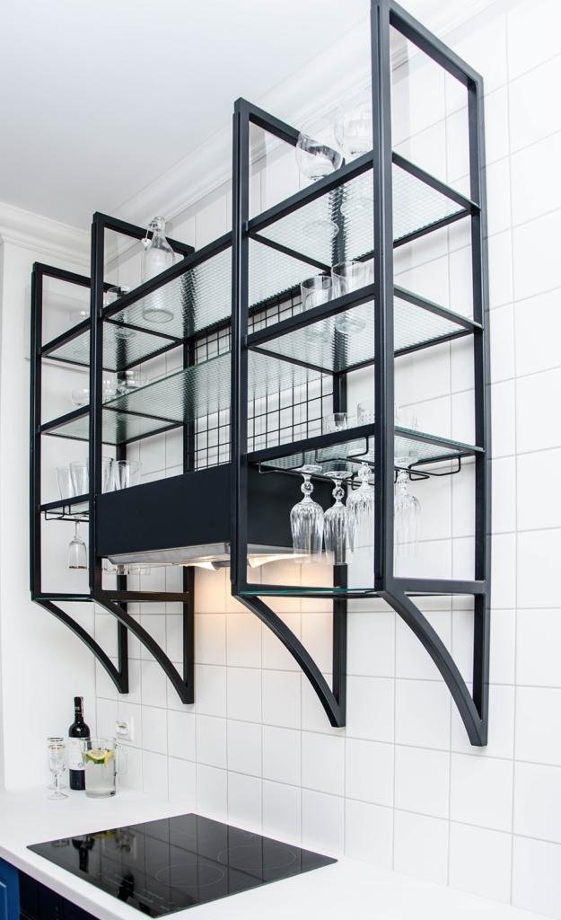 Kuchenny regał wiszący z czarnej stali z szklanymi półkami, podświetlony od dołu, na białej ścianie z płytek, z widoczną butelką wina i dzbankiem z napojem na blacie.