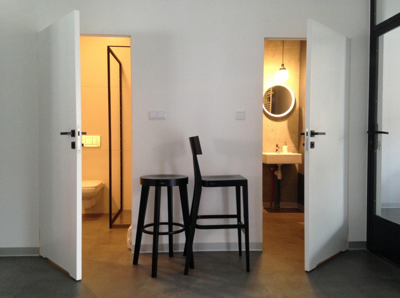 Minimalistyczne wnętrze z otwartymi drzwiami do łazienki i toalety, czarne krzesła barowe na pierwszym planie, dominująca biel i szarość, okrągłe lustro podświetlane w tle.