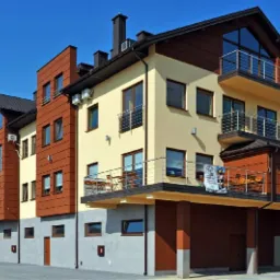 Nowoczesny, wielopiętrowy budynek z balkonami z metalowymi balustradami, wykończony w odcieniach beżu i brązu, z widoczną reklamą na jednym z balkonów.