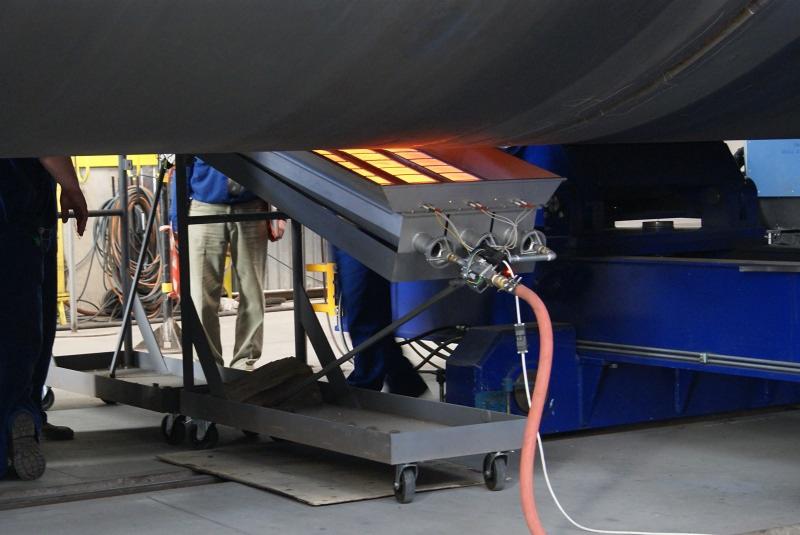 Przemysłowy promiennik ciepła na wózku podgrzewający rurę, widoczne palniki i przewody doprowadzające gaz.