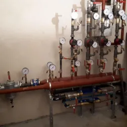 Rozbudowana instalacja hydrauliczna i grzewcza z licznymi manometrami, pompami obiegowymi i zaworami, zamontowana przy jasnej ścianie. Widoczne rury miedziane, stalowe i plastikowe, oraz elementy...