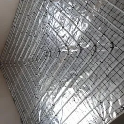 Białe rury ogrzewania podłogowego zamocowane na folii z nadrukowaną siatką, widoczne czarne uchwyty mocujące, surowy stan przed wylewką.