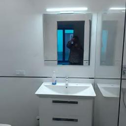 uslugi ogólno budowlane - Nowoczesna łazienka z białymi płytkami, umywalką z szafką i lustrem, w którym odbija się osoba robiąca zdjęcie.