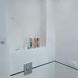 uslugi ogólno budowlane - Wykończona, minimalistyczna toaleta z białymi ścianami, szarymi akcentami fug i listew, oraz półką z akcesoriami montażowymi, w tym tubami silikonu i przyciskiem spłuczki WC w kolorze stali.