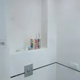 Wykończona, minimalistyczna toaleta z białymi ścianami, szarymi akcentami fug i listew, oraz półką z akcesoriami montażowymi, w tym tubami silikonu i przyciskiem spłuczki WC w kolorze stali.