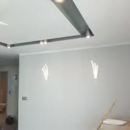 uslugi ogólno budowlane - Świeżo pomalowane na biało wnętrze z dekoracyjnymi lampami ściennymi w kształcie dłoni, widoczne narzędzia malarskie i zabezpieczone podłogi przed malowaniem.