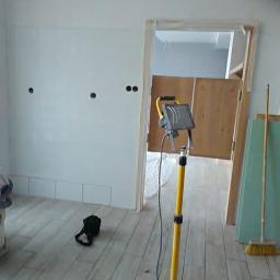 uslugi ogólno budowlane - Świeżo położona gładź na ścianie z widocznymi otworami na gniazdka, oświetlona halogenem budowlanym, z fragmentem płytek przypodłogowych i widoczną szczotką do zamiatania.