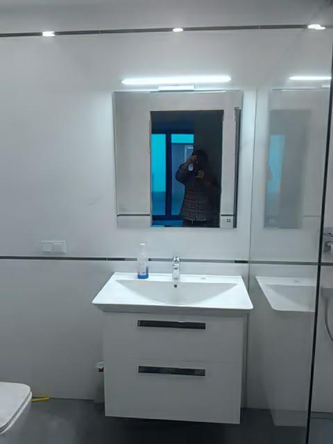 Nowoczesna łazienka z białymi płytkami, umywalką z szafką i lustrem, w którym odbija się osoba robiąca zdjęcie.