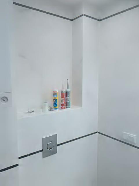 Wykończona, minimalistyczna toaleta z białymi ścianami, szarymi akcentami fug i listew, oraz półką z akcesoriami montażowymi, w tym tubami silikonu i przyciskiem spłuczki WC w kolorze stali.