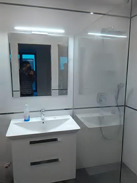 Nowoczesna łazienka z białą szafką pod umywalką, lustrem z oświetleniem LED i przeszkloną kabiną prysznicową z chromowaną baterią.