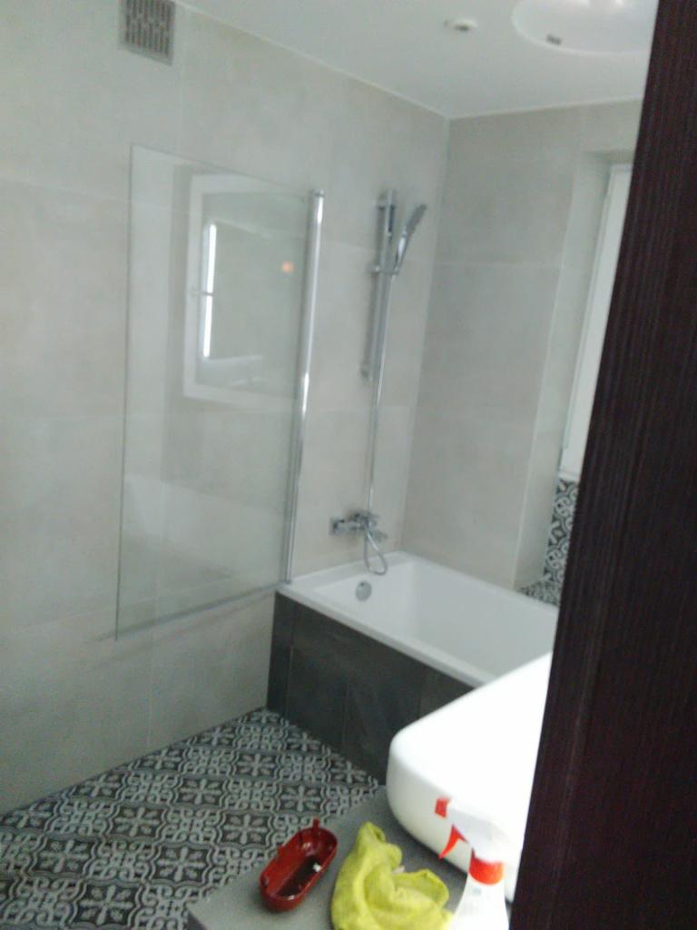 Nowoczesna łazienka z wanną obudowaną ciemnymi płytkami, szklanym panelem prysznicowym i wzorzystą podłogą. Widoczne akcesoria do czyszczenia na pierwszym planie.