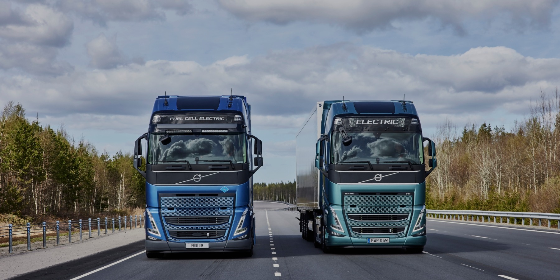 Dwa nowoczesne, elektryczne samochody ciężarowe Volvo: niebieski z napędem wodorowym i turkusowy, stojące na asfalcie drogi na tle lasu i pochmurnego nieba.