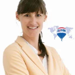 Uśmiechnięta kobieta w beżowej marynarce, z logo RE/MAX na tle mapy świata.