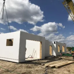 Montaż segmentów domu z prefabrykatów na fundamencie, widoczny dźwig budowlany i elementy konstrukcyjne.