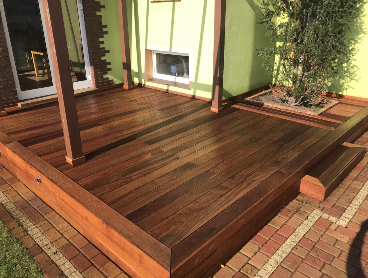 Nowoczesny taras z egzotycznego drewna o ciepłym, brązowym odcieniu, z niskimi stopniami wejściowymi i wbudowaną donicą dla drzewa, otoczony kostką brukową i zieloną ścianą budynku.