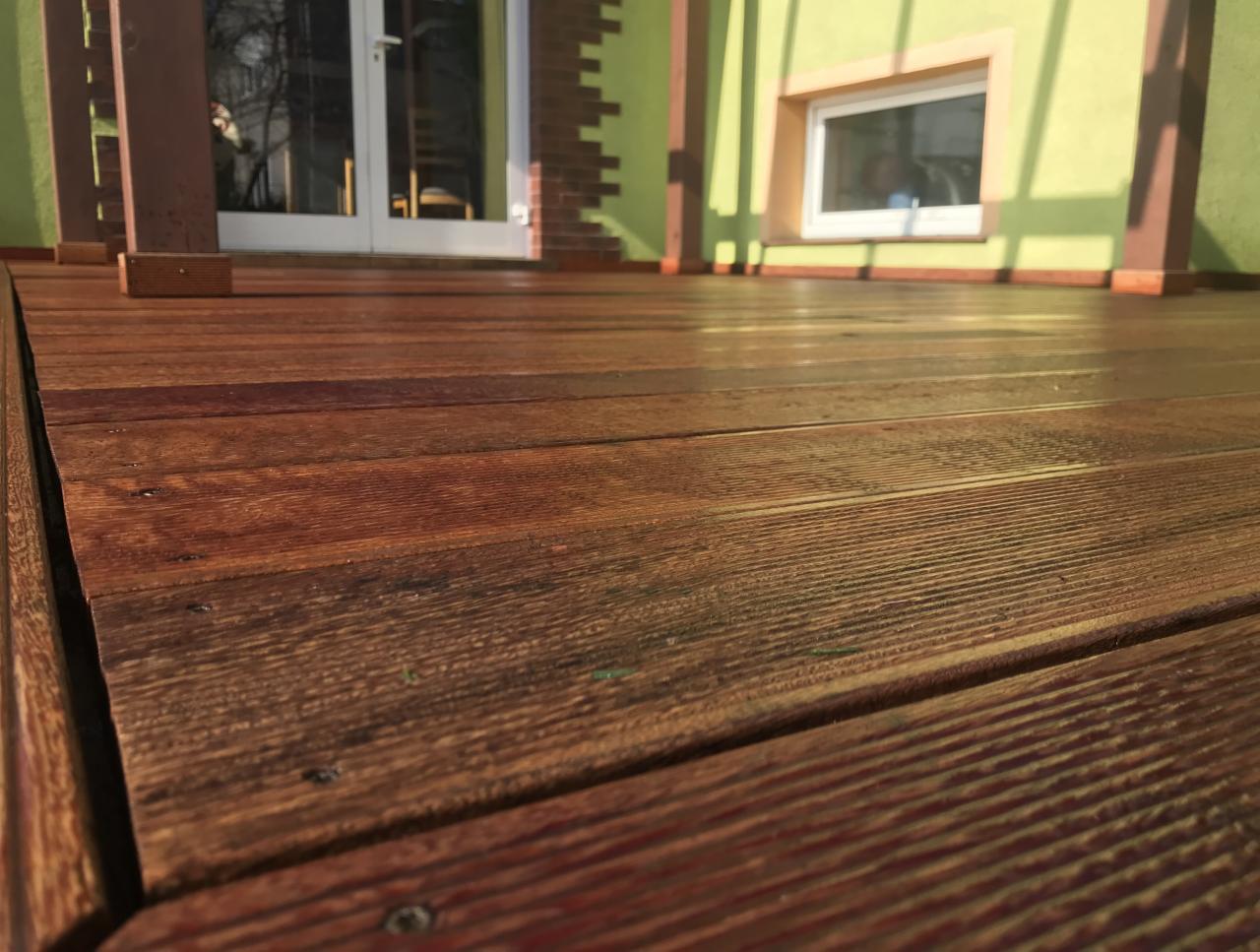 Błyszczący, brązowy taras drewniany z widocznymi śrubami, prowadzący do jasnego budynku z zieloną ścianą i białymi drzwiami.