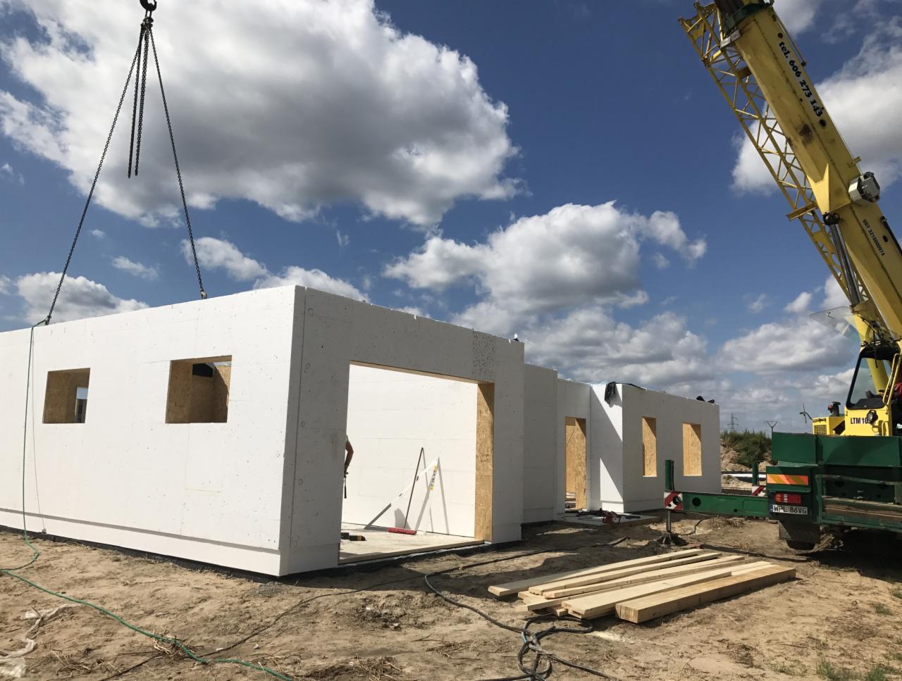 Montaż segmentów domu z prefabrykatów na fundamencie, widoczny dźwig budowlany i elementy konstrukcyjne.