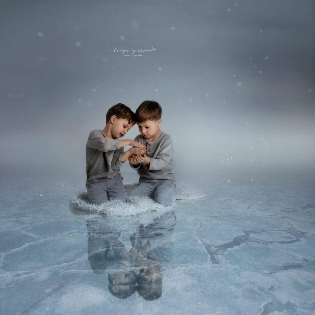 Dwóch chłopców w szarych ubraniach siedzi na białym futrze na tle imitującym lód i śnieg, z delikatnym efektem opadających płatków śniegu i odbiciem w lodowej tafli.