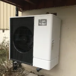 Biała jednostka zewnętrzna pompy ciepła Mitsubishi Electric Ecodan zamontowana na ścianie budynku, widoczne elementy mocujące i krzewy w tle.