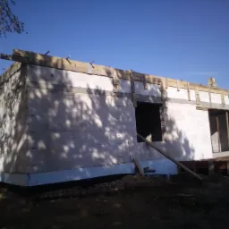 Surowy stan budynku z białych bloczków, widoczne drewniane elementy szalunku na górze ściany, ocieplenie fundamentów i zbrojenie w gruncie, cień drzewa na fasadzie, błękitne niebo w tle.