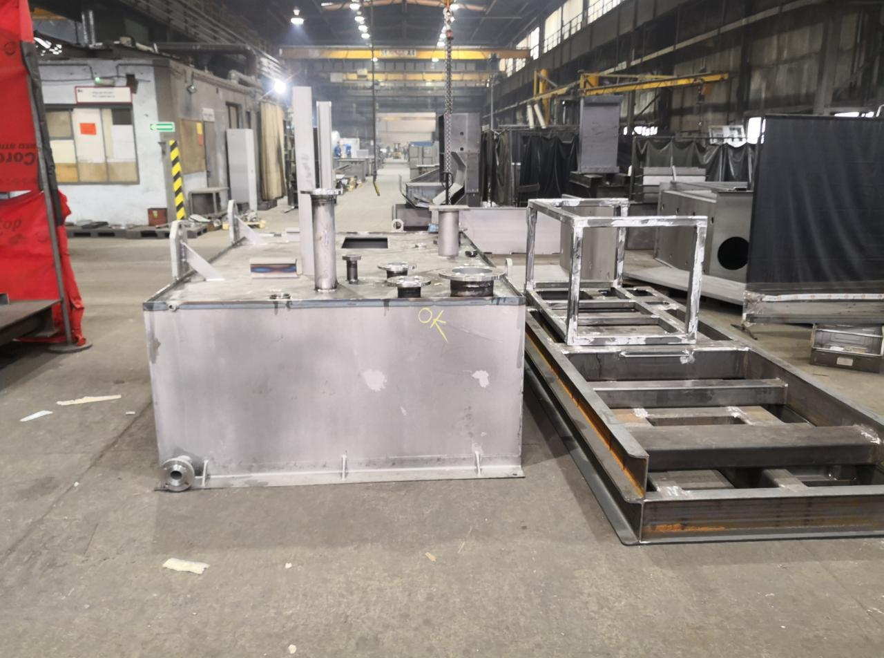 Wnętrze hali produkcyjnej z elementami konstrukcji stalowych: spawany zbiornik ze stali nierdzewnej z oznaczeniem 'OK' i fragment ramy, widoczne inne konstrukcje w tle.