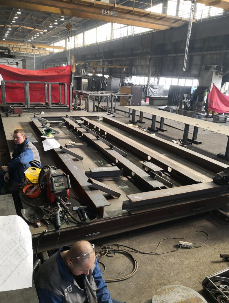 Wnętrze hali produkcyjnej: dwóch pracowników w kombinezonach przy konstrukcji stalowej, widoczny sprzęt spawalniczy, plany techniczne, suwnica pod sufitem.