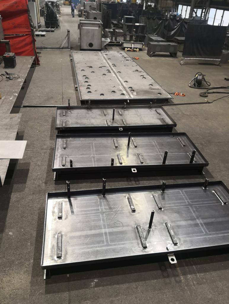 Metalowe płyty i konstrukcje w hali produkcyjnej, widoczne ślady obróbki i spawania, w tle inne elementy konstrukcyjne i osoba w oddali.