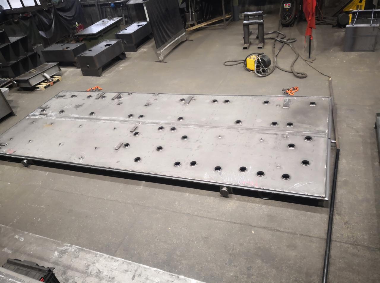 Duża, metalowa płyta z otworami i spawami w hali produkcyjnej, widoczny sprzęt spawalniczy w tle.