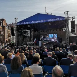 Uroczystość plenerowa z dużą sceną zadaszoną niebieskim baldachimem, na której widać przemawiającego mężczyznę przy mównicy, a za nim ekran LED i flagi, przed sceną zgromadzony tłum siedzący...