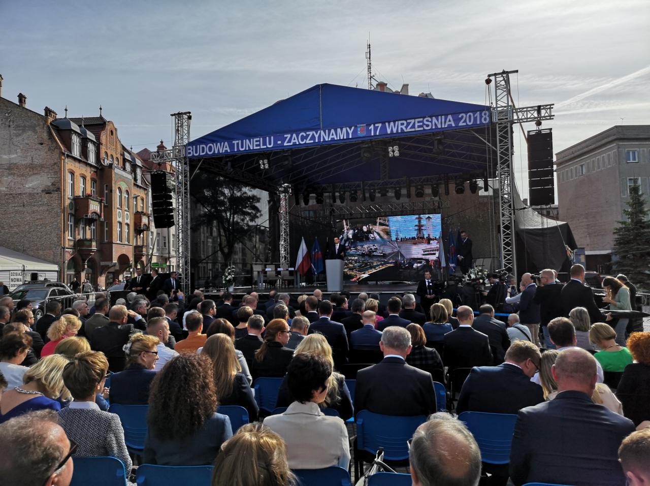Uroczystość plenerowa z dużą sceną zadaszoną niebieskim baldachimem, na której widać przemawiającego mężczyznę przy mównicy, a za nim ekran LED i flagi, przed sceną zgromadzony tłum siedzący...