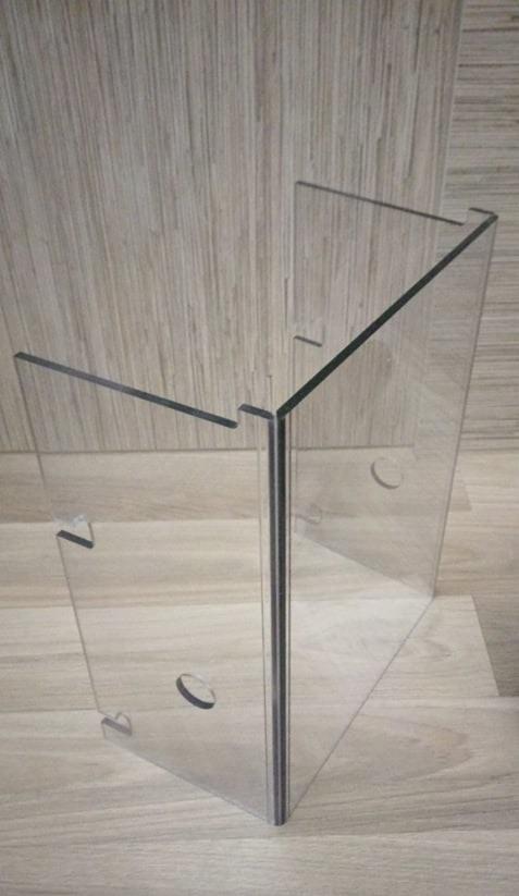 Kanciasta konstrukcja z przezroczystego akrylu z otworami, stojąca na drewnianej powierzchni i oparta o ścianę o podobnym wykończeniu.